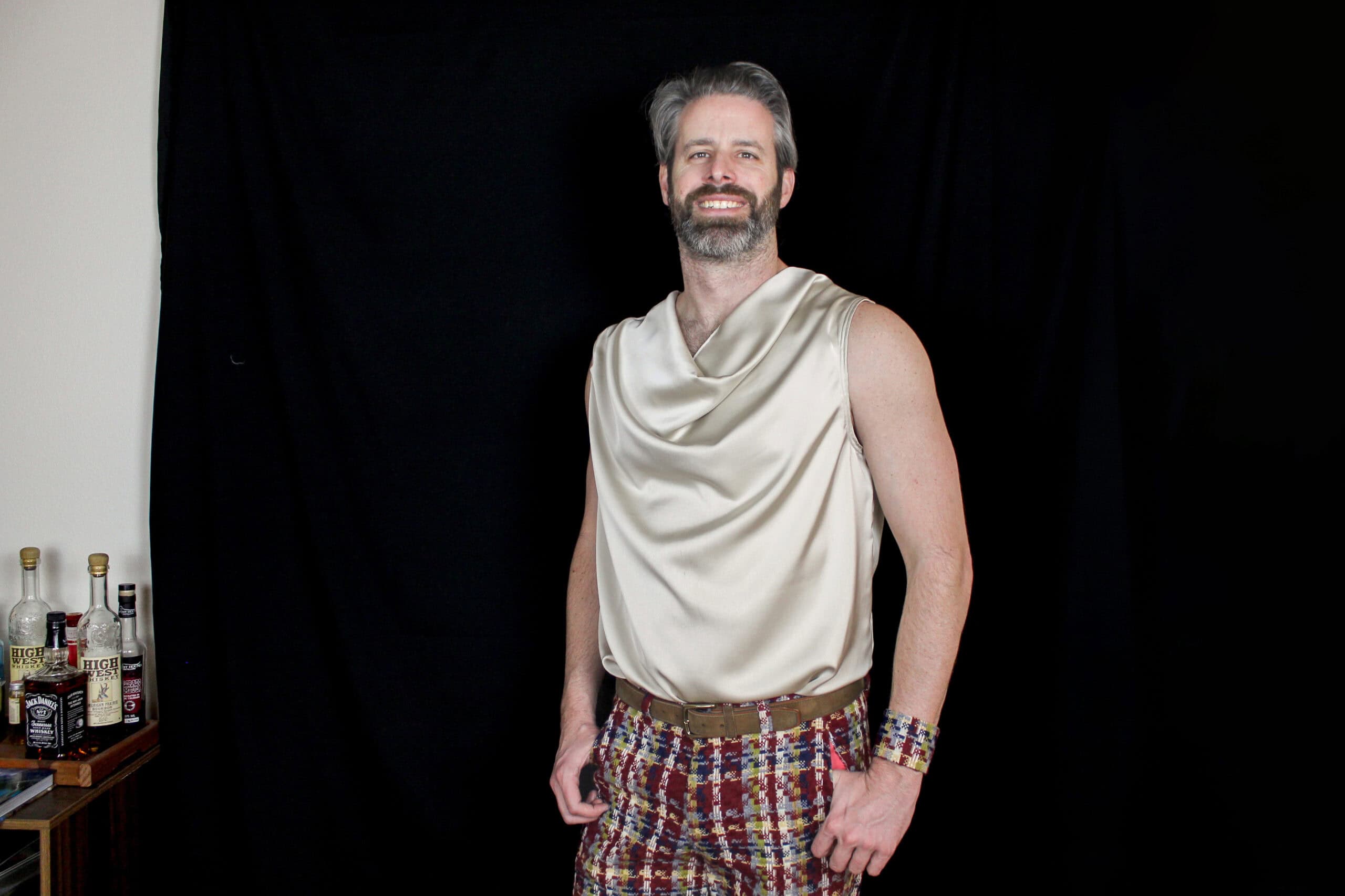 A man with a beard wearing a cream sleeveless top and colorful patterned pants, standing confidently.