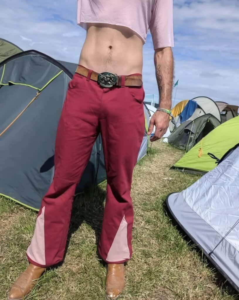 Sewist showcasing vibrant red and pink outdoor trousers with a belt, standing among tents at a camps.