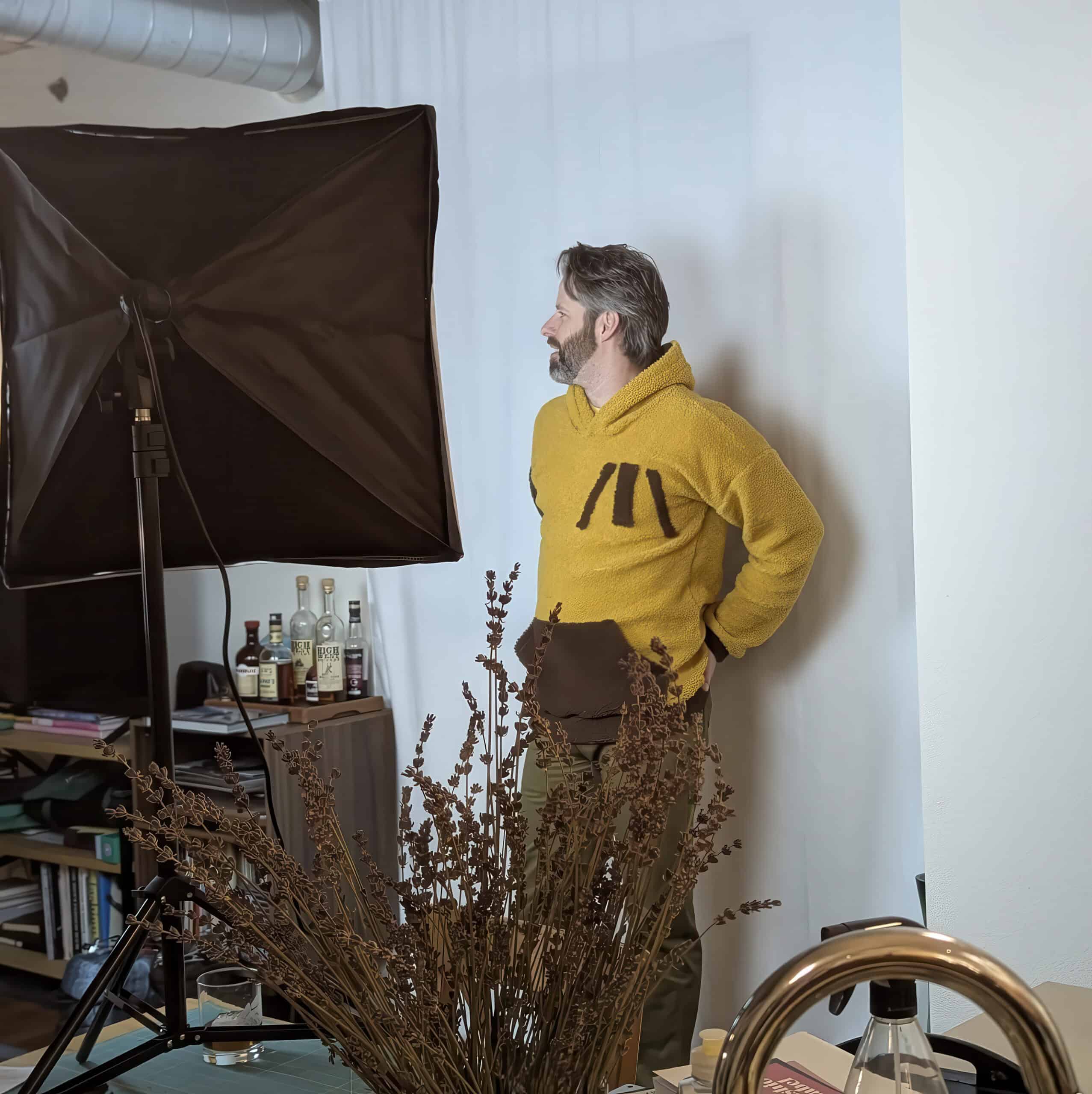 Man in yellow hoodie standing near photography setup and bottles.