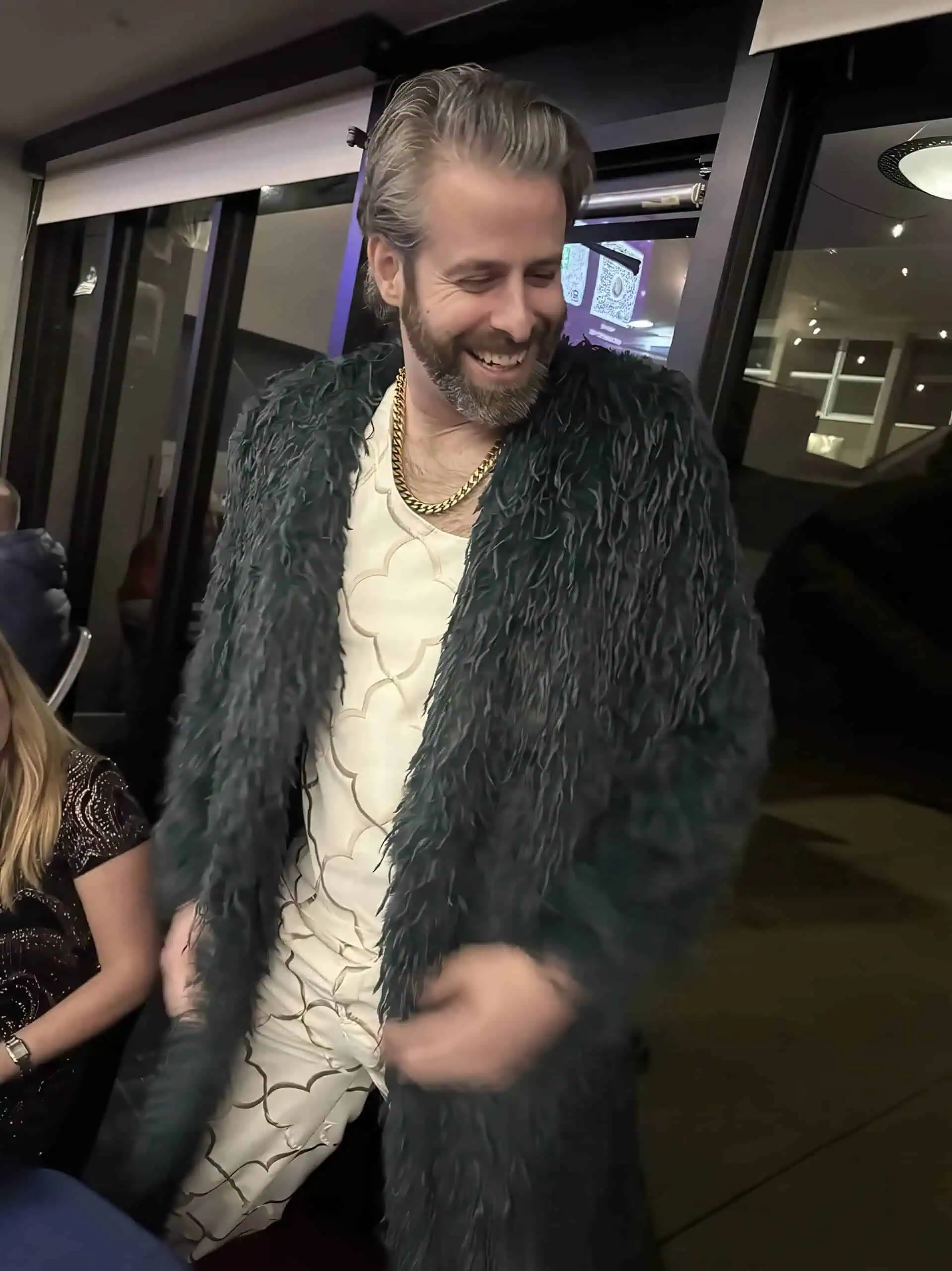 Man wearing a dark textured coat with a wide collar, cream patterned shirt, and gold chain necklace.