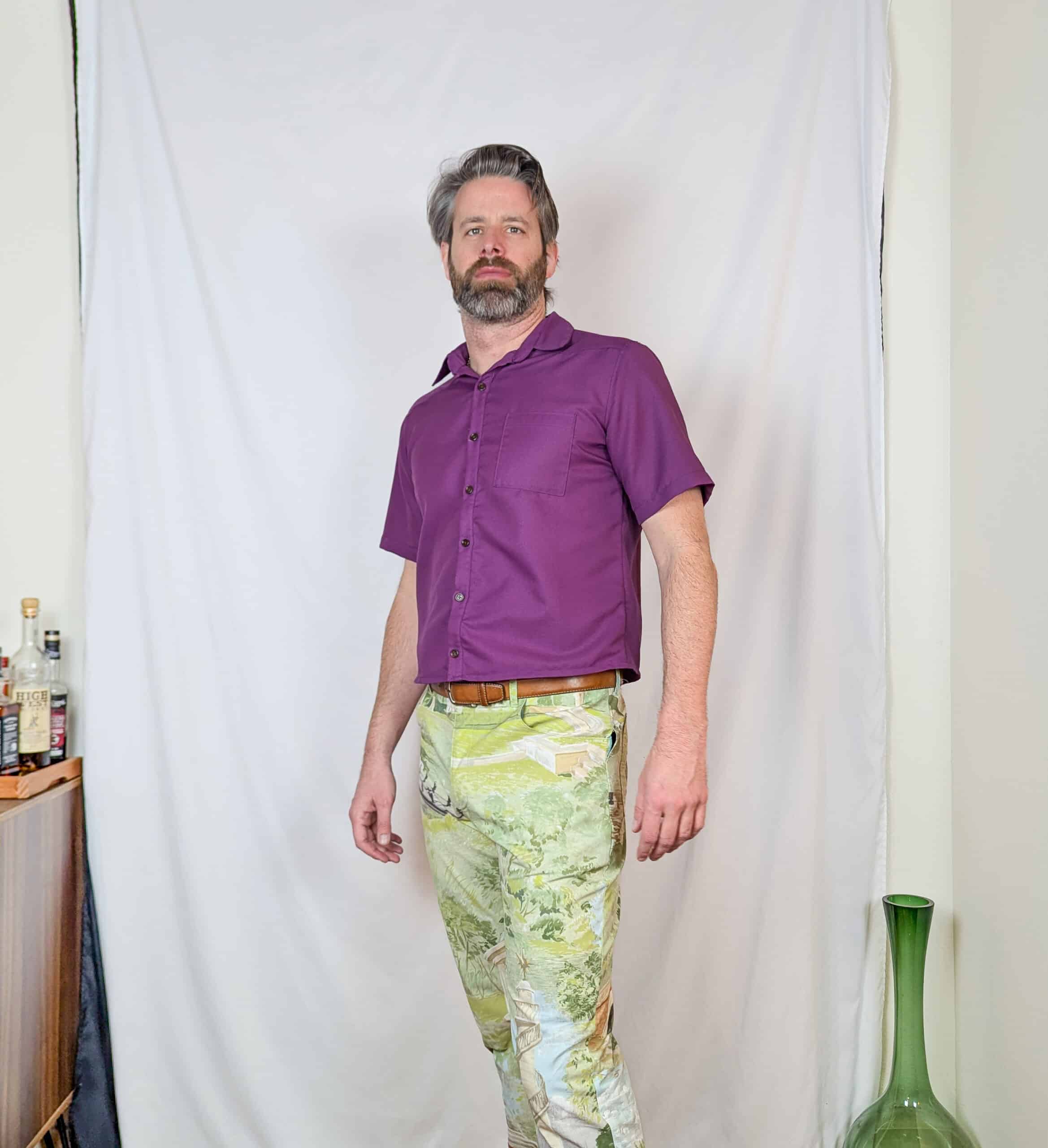 Male sewist wearing a purple short-sleeve shirt and colorful patterned pants, standing confidently i.